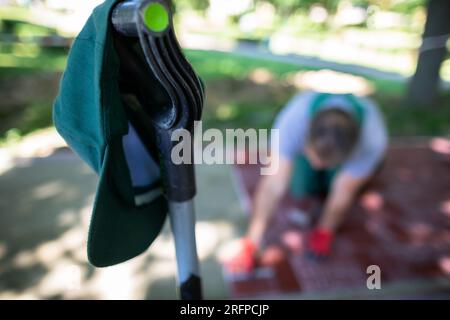 Eine grüne Baseballkappe hängt am Griff einer Schaufel, und im Hintergrund legt ein Arbeiter Pflastersteine. Stockfoto