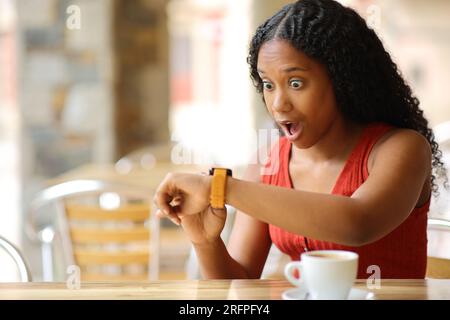 Überraschte schwarze Frau, die Smartwatch auf einer Barterrasse prüfte Stockfoto