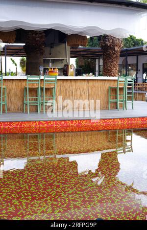 Tropic Bar am Pool an einem Sommertag mit Palmen im Hintergrund. Reflexion über Wasser und Stühle. Stockfoto