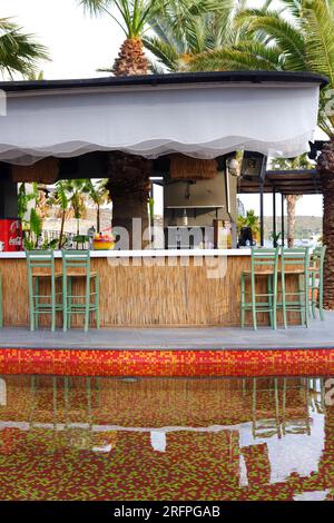 Tropic Bar am Pool an einem Sommertag mit Palmen im Hintergrund. Reflexion über Wasser und Stühle. Stockfoto