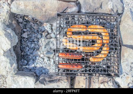 Würstchen und Kupaty werden auf einem Grill auf Kohlen zwischen Steinen gebraten Stockfoto