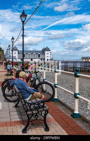 Ein Paar sitzt an einem Sommertag neben seinen Fahrrädern auf einer Strandpromenade. Pier im Hintergrund. Sonniger Tag. Entspannendes Konzept. Stockfoto