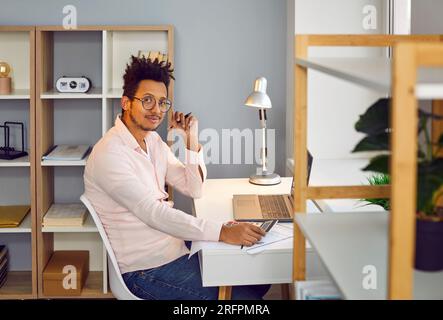 Ein junger Geschäftsmann, der am Schreibtisch im Büro arbeitet und lächelnd in die Kamera schaut. Stockfoto