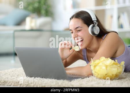 Lustige Frau, die Chips isst und sich zu Hause Online-Videos in einem Laptop ansieht Stockfoto