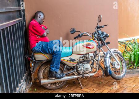 Ein boda-boda-Fahrer saß auf seinem Motorrad und lehnte sich an eine Wand. Er schlief im Unterschlupf einer Veranda ein und wartete auf den Sturm. Stockfoto