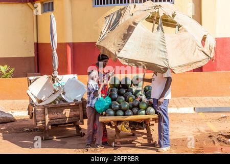 Geschützt unter einem Sonnenschirm verhandelt ein Wassermelonenverkäufer mit einer Frau und ihrem Sohn. Jinja, Uganda. Stockfoto