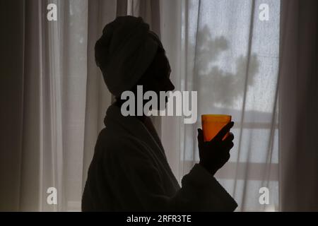 Junge schöne Frau in Handtüchern eingewickelt, die flüssiges Kollagen oder Vitaminergänzung neben dem Fenster trinken, gesundes Morgen-Lifestyle-Konzept Stockfoto