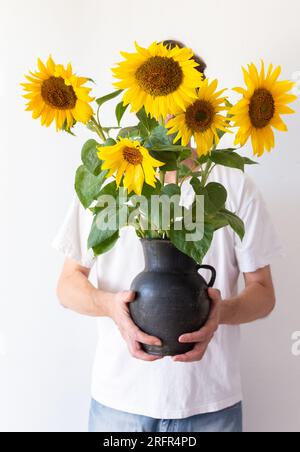 Ein Mann hält einen schwarzen Krug mit Sonnenblumen vor sich an eine weiße Wand Stockfoto