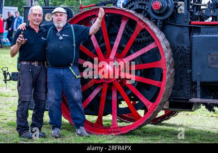 Alt Schwerin, Deutschland. 05. Aug. 2023. Holger Severin (l) und Maik Lemke stehen vor dem Antriebsrad einer zehn Tonnen schweren „MAN“-Lokomotive, die 1918 auf der diesjährigen Dampfkonferenz im Agroneum Museum of Agricultural History gebaut wurde. Vor allem Technologiefans aus den Niederlanden, die wegen Corona nicht zum letzten Treffen vor zwei Jahren reisen konnten, sind wieder in das Mecklenburg-Seengebiet gereist. Insgesamt sind mehr als 50 dampfbetriebene Traktoren und Maschinen in Aktion zu sehen, darunter etwa 13 große Maschinen. Kredit: Jens Büttner/dpa/Alamy Live News Stockfoto