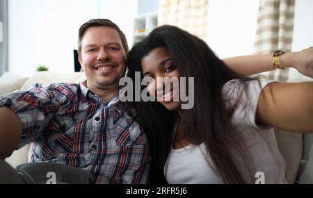 Portrait eines fröhlichen gemischten Paares, das fröhliches Selfie auf dem Sofa drinnen macht. Stockfoto