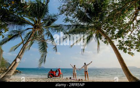 Motorroller Road Trip. Schönes Paar auf rotem Motorrad in weißen Kleidern am Sandstrand. Gerade verheiratete Menschen küssen Umarmungen, die in der Nähe der tropischen Palmen, dem Meer, spazieren gehen. Hochzeitsreise am Meer. Motorradmiete. Stockfoto