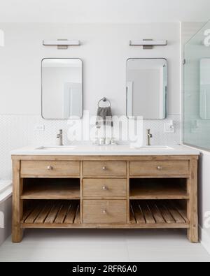 Ein gemütliches Badezimmer mit Schrank aus weißer Eiche, weißer Arbeitsfläche, bronzefarbenen Wasserhähnen und Lampen und weißen Fischgrätfliesen im Hintergrund. Stockfoto