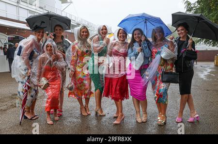 Rennfahrer, die Ponchos tragen und Regenschirme halten, um sich vor Regen zu schützen, am 5. Tag des Qatar Goodwood Festivals 2023 auf der Rennbahn Goodwood, Chichester. Foto: Samstag, 5. August 2023. Stockfoto