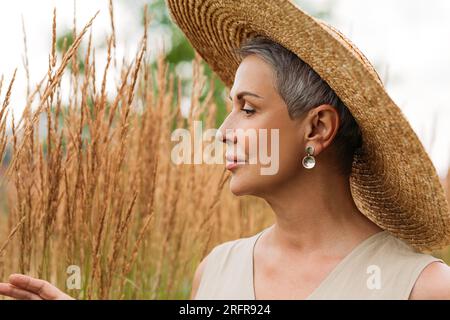 Seitenansicht einer Seniorin mit Strohhut, die Weizen auf einem Feld ansieht Stockfoto