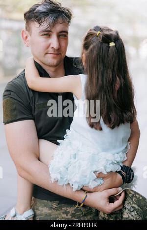 Portrait eines glücklichen ukrainischen Soldaten, der aus dem Krieg zurückkehrt, seine kleine Tochter trifft und sie umarmt. Krieg in der Ukraine. Russische Militärinvasion i Stockfoto