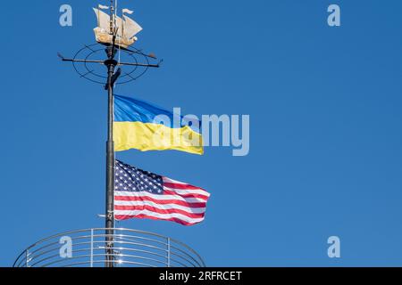 Die Flaggen der Ukraine und der USA flattern auf einem Fahnenmast vor einem blauen Himmel. Statussymbol. Ukrainische blaue und gelbe Flagge. Windtag im Freien. Stockfoto