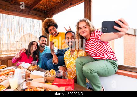 Eine multiethnische Gruppe glücklicher junger Freunde veranstaltet zu Hause eine Barbecue-Dinner-Party auf dem Dach - multiethnische, fröhliche junge Erwachsene, die Spaß und Bindung haben Stockfoto