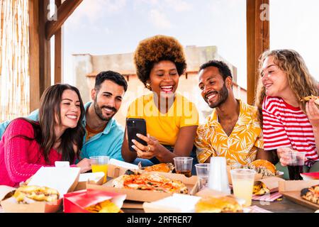 Eine multiethnische Gruppe glücklicher junger Freunde veranstaltet zu Hause eine Barbecue-Dinner-Party auf dem Dach - multiethnische, fröhliche junge Erwachsene, die Spaß und Bindung haben Stockfoto