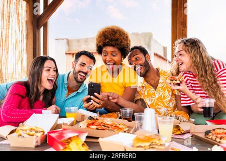 Eine multiethnische Gruppe glücklicher junger Freunde veranstaltet zu Hause eine Barbecue-Dinner-Party auf dem Dach - multiethnische, fröhliche junge Erwachsene, die Spaß und Bindung haben Stockfoto