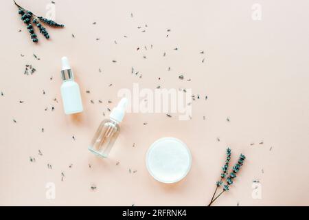 Lavendel, natürliches Kosmetikserum, ätherisches Öl und Cremebecher auf pastellbeigefarbenem Hintergrund mit Blumen. Aromatherapie, Hautpflegekosmetikkonzept. Oben vi Stockfoto