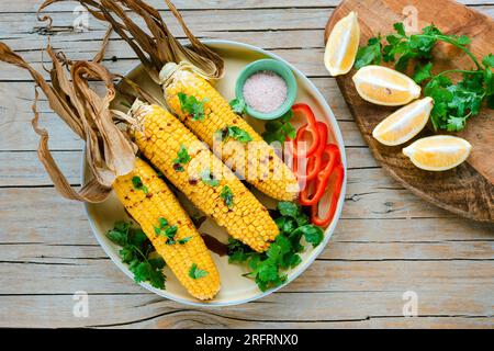 Gebackene Maiskolben mit Koriander, in Scheiben geschnittenem Paprika, Zitrone und Salz auf einem Holztisch. Mexikanisches Speisekonzept. Draufsicht, flach liegend. Stockfoto