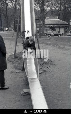 1969, historisch, ein kleiner Junge, der auf einem traditionellen Metallspielplatz zuerst auf dem Bauch runterrutscht, England, Großbritannien. Stockfoto