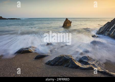 Küstenfelsen, die bei Sonnenuntergang von Wellen bei aufsteigender Flut gewaschen werden; Ayrmer Cove, bei Kingsbridge, an der Südküste von Devon, Großbritannien. Stockfoto