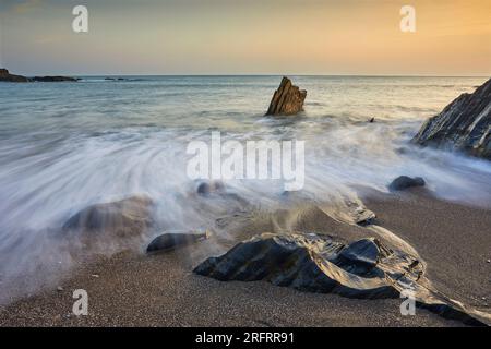 Küstenfelsen, die bei Sonnenuntergang von Wellen bei aufsteigender Flut gewaschen werden; Ayrmer Cove, bei Kingsbridge, an der Südküste von Devon, Großbritannien. Stockfoto