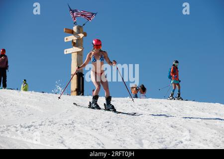 Mammoth Lakes, Kalifornien. 4. Juli 2023. An klaren Sommertagen kommt eine fitte Frau in einem Bikini und Helm vorbei an amerikanischen Flaggen im Mammoth Mountain Ski Resort. Stockfoto