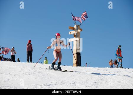 Mammoth Lakes, Kalifornien. 4. Juli 2023. An klaren Sommertagen kommt eine fitte Frau in einem Bikini und Helm vorbei an amerikanischen Flaggen im Mammoth Mountain Ski Resort. Stockfoto