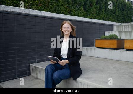 Eine lächelnde blonde Frau mit trendiger Sonnenbrille geht die Straße entlang und benutzt ihr Telefon. Stockfoto