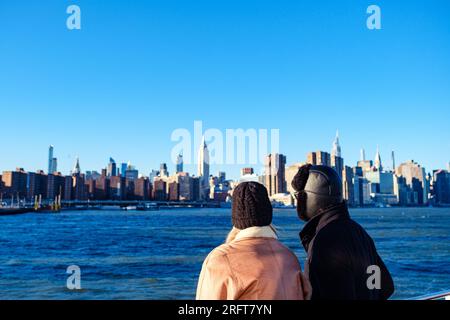 Sonnendurchflutete Paare in Winterkleidung, die das pulsierende Midtown Manhattan vom East River aus betrachten Stockfoto