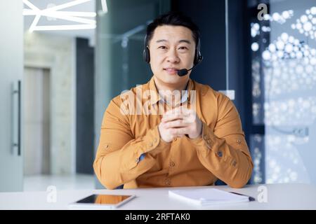 Porträt eines Geschäftsmanns, der im Kundendienst für Online-Käufer tätig ist, ein Mann am Arbeitsplatz, der lächelt und in die Kamera schaut, ein asiatischer Mann mit Headset für Videoanrufe. Stockfoto