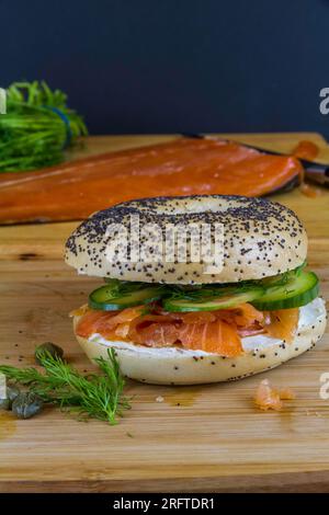 Bagel mit Räucherlachs, Frischkäse, Gurke, Dill und Kapern auf Holzbrett, Halbseite im Hintergrund, Hochformat. Stockfoto