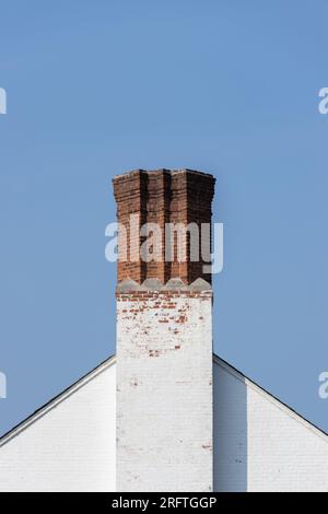 Ein einzigartiger Kamin aus rotem Ziegelstein mit geometrischen Winkeln auf einem weißen Ziegeldach vor einem klaren blauen Himmel, Kopierraum Stockfoto