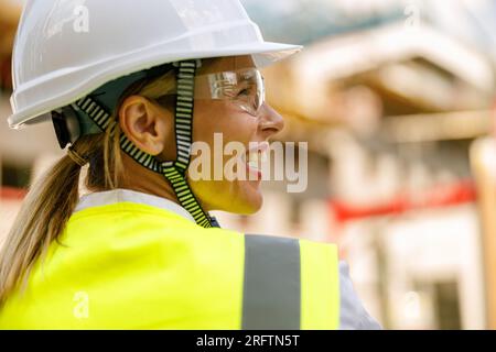 Lächelnde Bauarbeiterin im Schutzhelm, die auf dem Bauhintergrund steht Stockfoto