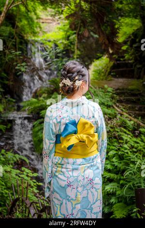 Rückansicht einer Frau mit japanischem Yukata-Sommerkimono, die auf der Straße neben dem Waldwasserfall in der Natur spaziert. Kyoto, Japan. Stockfoto