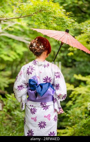 Rückansicht einer Frau, die einen japanischen Yukata-Sommerkimono trägt und einen traditionellen japanischen Ölpapierschirm im Naturwald hält. Kyoto, Japan. Stockfoto