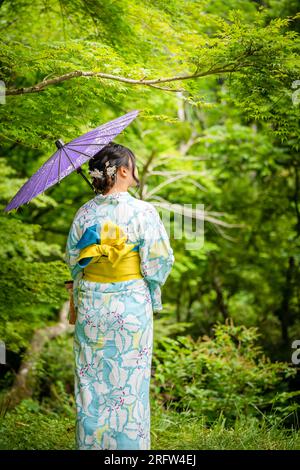 Rückansicht einer Frau, die einen japanischen Yukata-Sommerkimono trägt und einen traditionellen japanischen Ölpapierschirm im Naturwald hält. Kyoto, Japan. Stockfoto