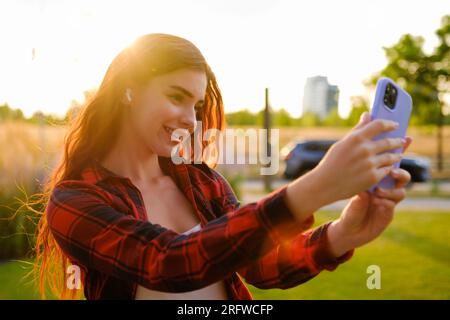 Ein rothaariges Mädchen, das ein rot-schwarzes Hemd trägt, macht bei Sonnenuntergang ein Selfie mit einem Smartphone. Stockfoto