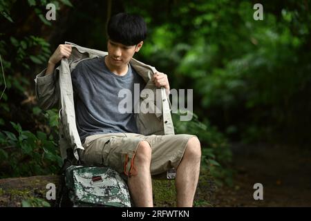 Ein asiatischer Mann trägt Campingkleidung und Rucksack, der auf einem Baumstamm auf der Straße ruht, während er die Natur erkundet. Stockfoto