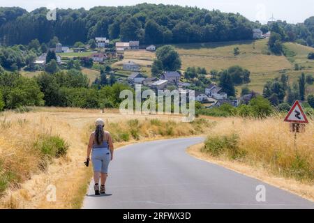 Asphaltstraße auf dem Land mit erwachsenen weiblichen Wanderinnen auf der linken Seite, deutscher Landschaft, Dorf Utscheid und grünen grünen Bäumen auf einem Hügel im Hintergrund, c Stockfoto