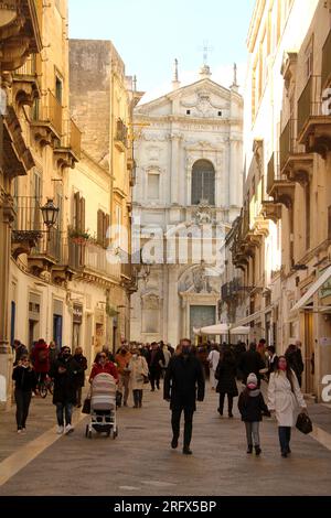 Lecce, Italien. Menschen, die während der COVID-19-Pandemie auf der Via Vittorio Emanuele spazieren gehen und Masken tragen. Die Kirche der Heiligen Irene hinten. Stockfoto