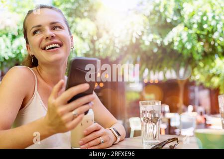 Glückliche junge Frau, die im Sommer im Straßencafé mit ihrem Handy saß. Stockfoto
