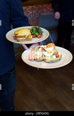 Der Kellner bringt Frühstück, zwei Gerichte, klassischer französischer Toast mit Käse und Schinken und Avocado-Toast mit weich gekochten Eiern Stockfoto