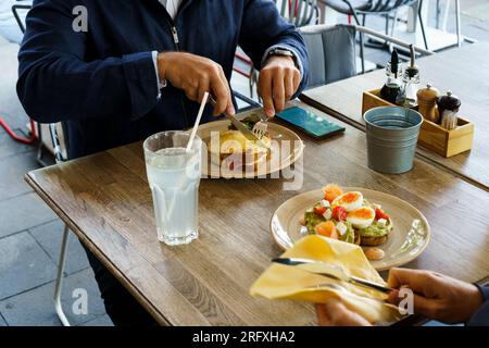 Zwei Personen essen Frühstücksgerichte, klassischer französischer Toast mit Käse und Schinken und Avocado-Toast mit weich gekochten Eiern Stockfoto