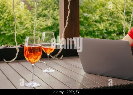 Laptop mit einem Glas Aperol-Cocktail. Unbekannte Person, die an einem drahtlosen Computer im Café im Freien arbeitet. Freiberufler in der Öffentlichkeit. Laptop und zwei Gläser Stockfoto