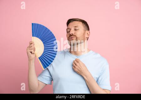 Das Porträt eines verspielten, müden Mannes, der mit einem blauen Ventilator wedelt, leidet an Stuffigkeit, wenn er auf pinkfarbenem Hintergrund steht. Stockfoto