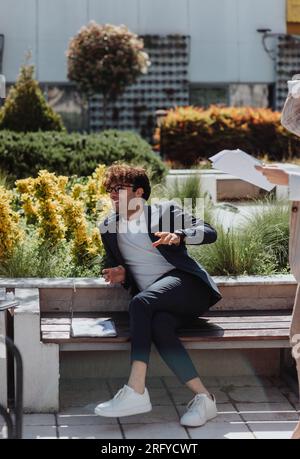 Geschäftsleute, die Remote arbeiten. Männlicher Geschäftsmann, der mit seinen Teamkollegen spricht, während er auf einer Bank in der Stadt liegt Stockfoto