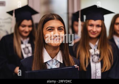 Junge Frau, die ihren Abschluss gemacht hat, voller Freude und Glück, lächelt und schaut in die Kamera für ihr Jahrbuchfoto mit den anderen Schülern im Hintergrund Stockfoto